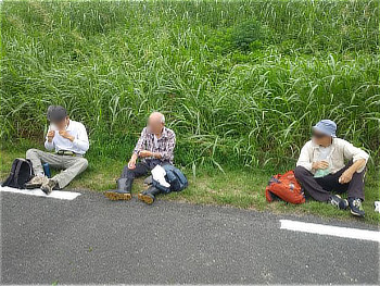 舗装道路の脇の草むら前に座り、休憩をとる3人の作業参加者