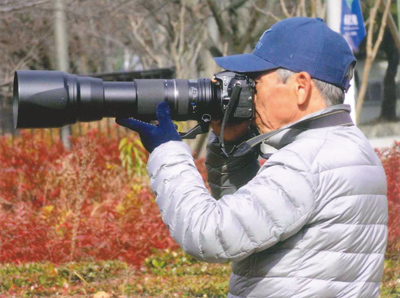 鳥たちの一瞬を狙ってシャッターをきる。(カメラを構える村上さんの姿の写真)