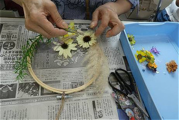 新聞紙を敷いたテーブルで、手を使って刺繍枠にドライフラワーを配置し、リースを作っている手元の様子。