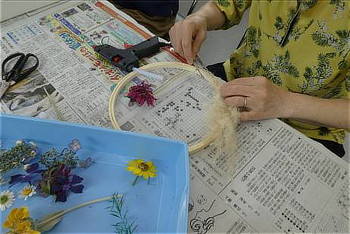 新聞紙を敷いたテーブルで、グルーガンを使い、刺繍枠にドライフラワーを接着してリースを作っている手元の様子。