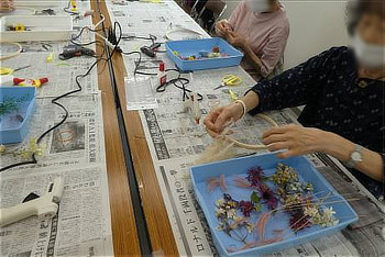 新聞紙が敷かれたテーブルに、水色のトレイに入った花材、グルーガン、刺繍枠が置かれ、参加者がリースを手作りしている風景。