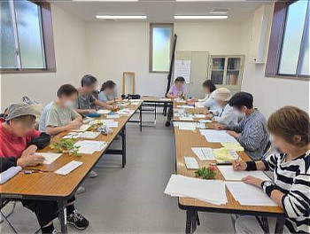 明るい会議室で長机を囲み、手元の木の実を見つめながら紙に向かってデッサンや彩色に集中するボタニカルアート体験講座の参加者たち
